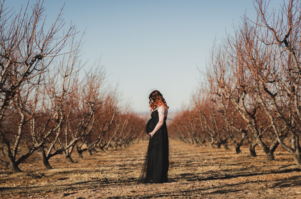 Dark and moody maternity photoshoot shot in llano california at a peach orchard. All the leaves have turned orange. The pregnant woman is wearing a black dress and heels and wearing a fox pelt on her head