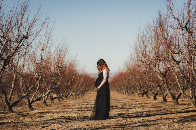 Dark and moody maternity photoshoot shot in llano california at a peach orchard. All the leaves have turned orange. The pregnant woman is wearing a black dress and heels and wearing a fox pelt on her head