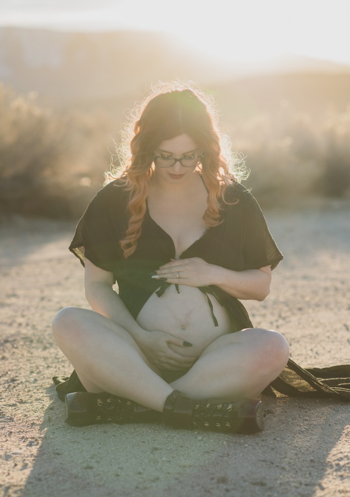 Dark and moody maternity photoshoot shot in llano california at a peach orchard. All the leaves have turned orange. The pregnant woman is wearing a black dress and heels and wearing a fox pelt on her head