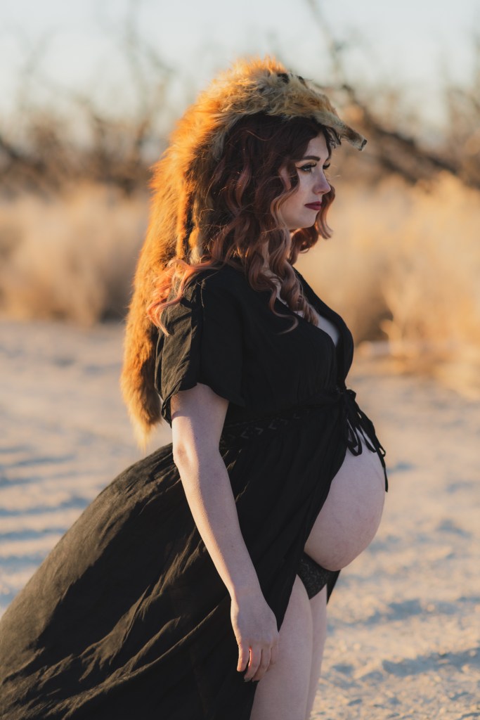 Dark and moody maternity photoshoot shot in llano california at a peach orchard. All the leaves have turned orange. The pregnant woman is wearing a black dress and heels and wearing a fox pelt on her head