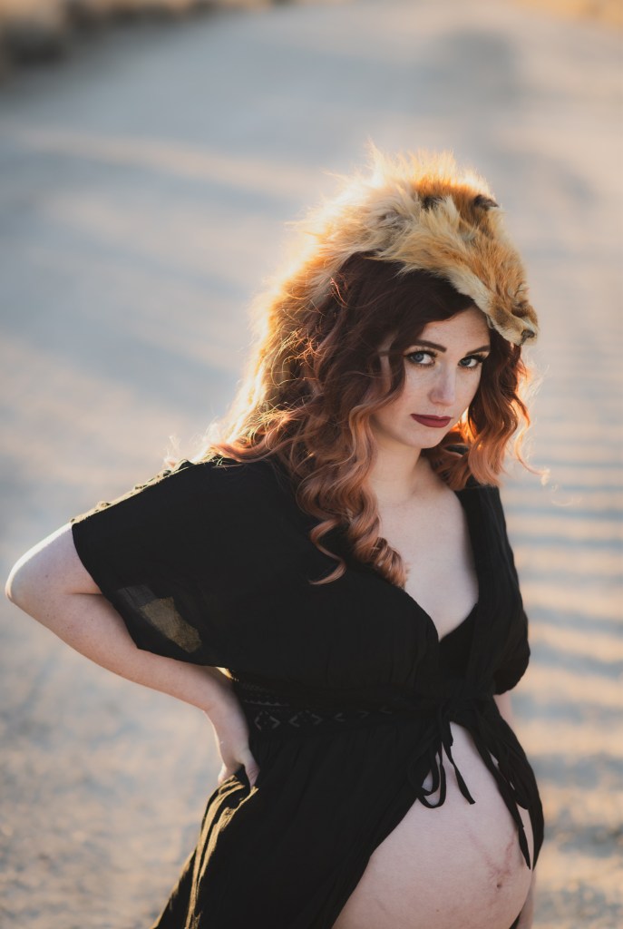 Dark and moody maternity photoshoot shot in llano california at a peach orchard. All the leaves have turned orange. The pregnant woman is wearing a black dress and heels and wearing a fox pelt on her head