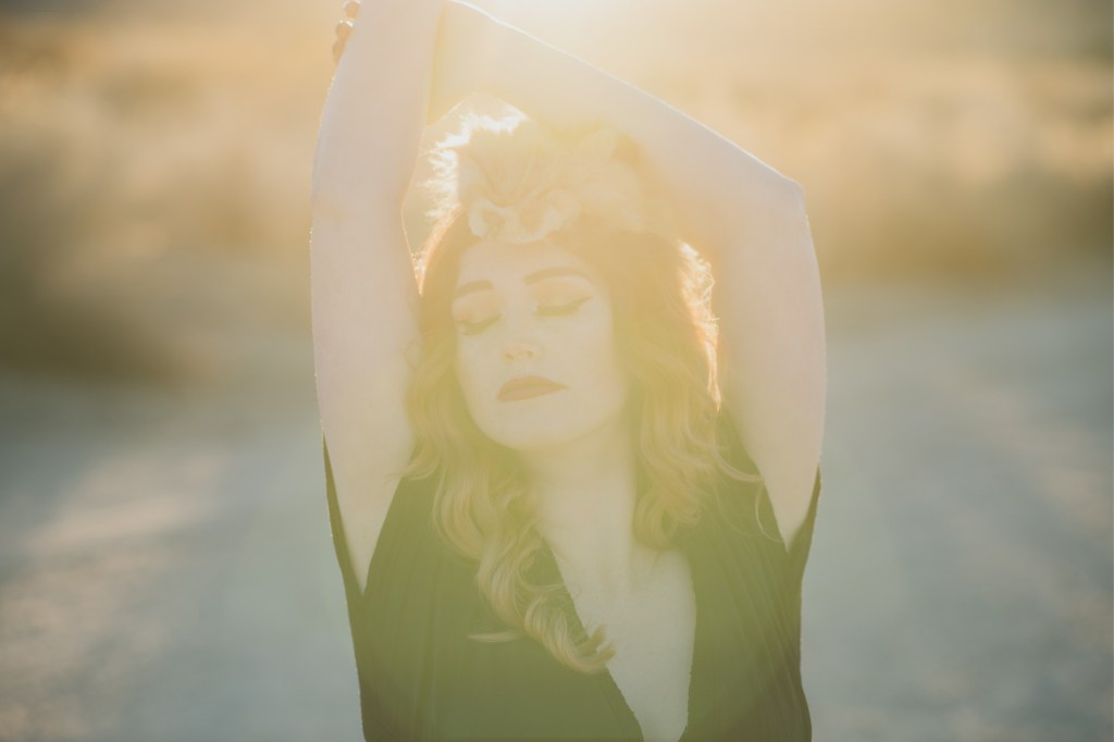 Dark and moody maternity photoshoot shot in llano california at a peach orchard. All the leaves have turned orange. The pregnant woman is wearing a black dress and heels and wearing a fox pelt on her head