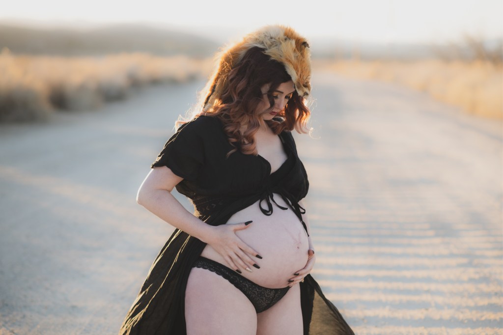 Dark and moody maternity photoshoot shot in llano california at a peach orchard. All the leaves have turned orange. The pregnant woman is wearing a black dress and heels and wearing a fox pelt on her head